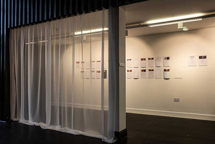 A4 sized artworks are displayed on a white wall. There is a see through curtain covering one of the archways into the space.