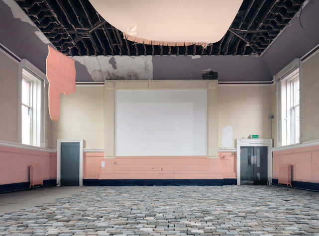 Installation view of the interior of a large room with a cobbled floor and a projection screen on the back wall