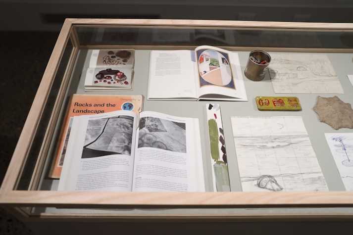 Open books and pencil drawings sit in a glass vitrine. One book is titled 'Rocks and the Landscape'.