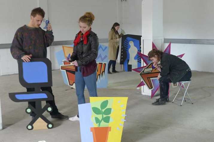 4 people in a room amongst 2d cartoony sculptures such as of a desk chair and a potted plant.