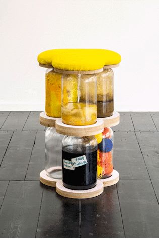 The image shows a sculpture of 6 large glass jars stacked and filled with materials