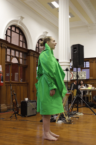 A woman dressed in a bright long coat, is standing barefoot and speaking at something ahead of her. There is a speaker placed behind her, behind which, there is a sign stating "Glasgow Women's Library".