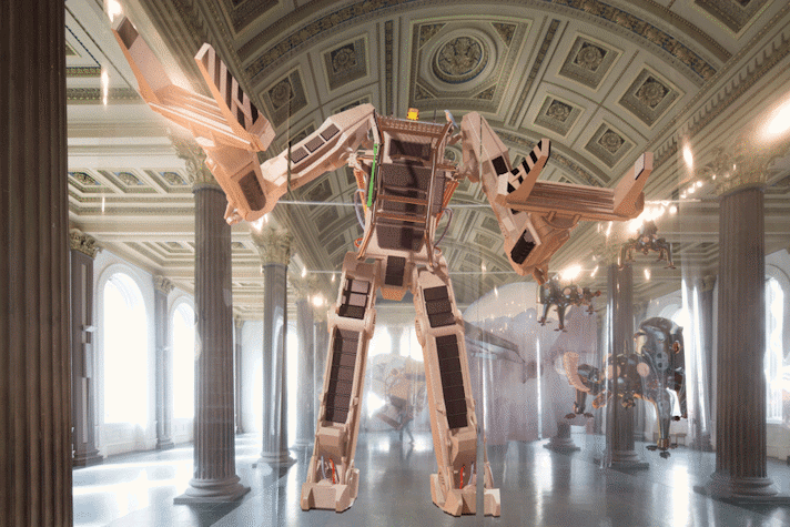 A giant robot is printed onto suspended panels of polyester foil. The transformer like robot has clamps for hands. There are two different coloured wires running through the legs and body of the robot, with a small bright light in place of the head.