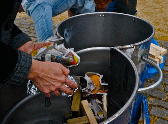 A pair of hands are holding some garbage over a couple joint trash cans. There is dry garbage in the trash cans, including newspaper and orange panels. There is another person behind the trash cans, who is picking up something from the ground.