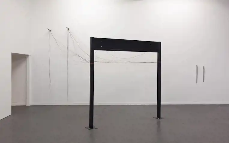 A large black metal structure of a beam with two legs stands in the space. Multi coloured strings form a messy net between the structure and two small abstract aluminium shapes on the wall.