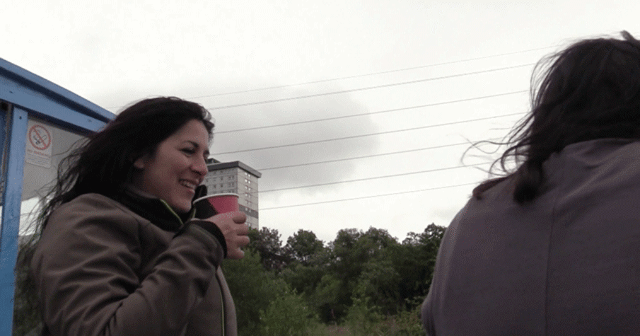 Two people with long brown hair stand in the wind one is holding a paper cup and is smiling