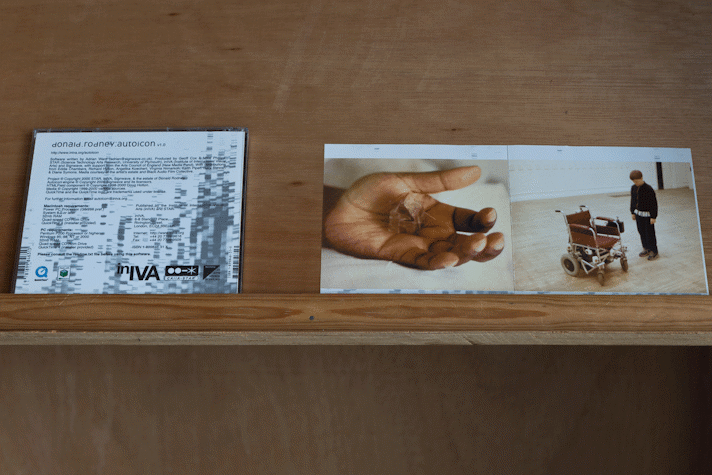 On a ply shelf there is a CD case with black and white sleeve that reads 'donald rodney: auto icon' and smaller black text with software and production credits. To the right of the CD case are two square images side by side. One shows a black person's hand, in their palm is what appears to be a small house like structure. The other image shows a wheelchair with brown leather seat, back and armrests and a light-skinned person with brown hair in a black jacket looking down at the chair.