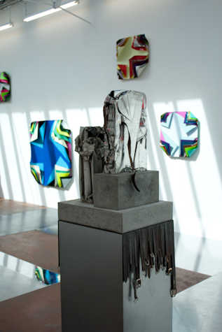 metal sculpture atop a plinth. from the plinth multiple black leather belts are hanging. behind there are sculptural works mounted on the wall of sheets folding in at the corners revealing more and more layers.