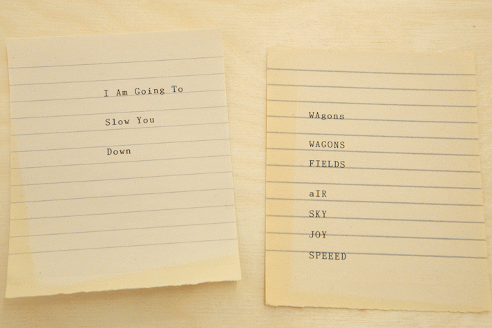 Placed on a discoloured and old sheet of taper are two ruled sheets with poems on them. These sheets are also old and discoloured. The one on the left reads, "I Am Going To, Slow You, Down" and the one on the right reads "Wagons: Wagons, Fields. Air, Sky, Joy, Speed."