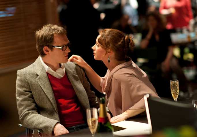 man and a woman sat in front of an audience. the woman has her hand on the mans shoulder and leans towards him. There are two glasses of champagne on the table in front of them.
