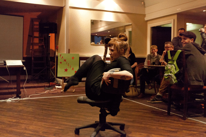 performance by Rose Ruane. The performer sits on spinning desk chair with their knees up as the audience watches.