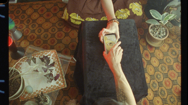 a photograph taken from above showing two people sitting on opposites sides of a black table.  Their arms reach towards the centre of the table and between them they hold an image of an eye. In the room surrounding them sits some potted plants and two lava lamps
