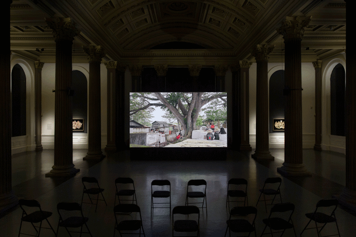 installation view of the GoMA. it is dimly lit and surrounded by pillars and arches is a projection screen showing a tree in a village,