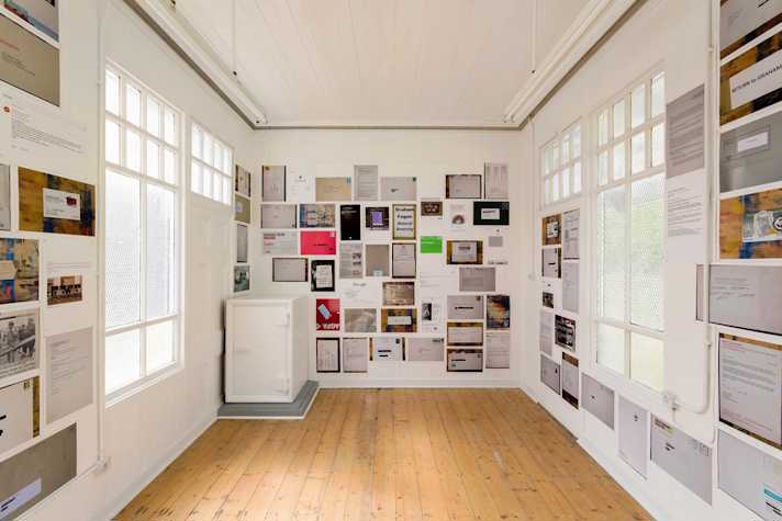 colourful photocopied sheets are pasted to the walls of a small gallery space. The sheets show letters, emails documents and other text-based media.
