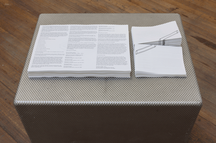 On a wooden floor there is a metallic, textured box. The box has a round dent like pattern all over it. On top of the box, there are leaflets and brochures piled up next to each other.