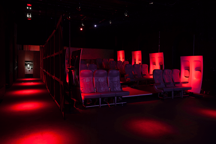 Image of Jenkin van Zyl's installation at Tramway. The room sits in eerie red light. There are aeroplane seats laid out and aeroplane window panels along the wall.