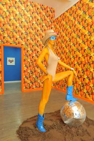 orange mannequin wearing a swimsuit, bright blue cowboy boots, a blue eye mask and a straw hat. the mannequin stands with one hand on its hips and its foot resting atop a disco ball. beneath the disco ball there is a pile of dirt. the room is wallpapered in a bright orange and blue pattern