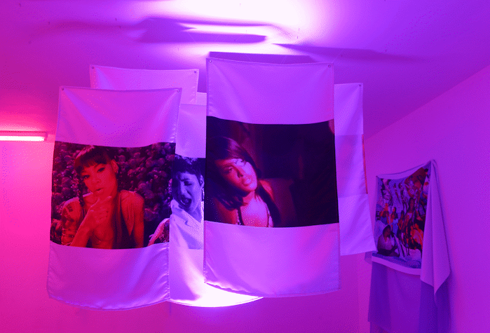 A room with pink neon lights has four fabrics hanging from the ceiling with images of women on it as a part of E. Jane's installation in Kelvin Hall as part of Glasgow International 2018.