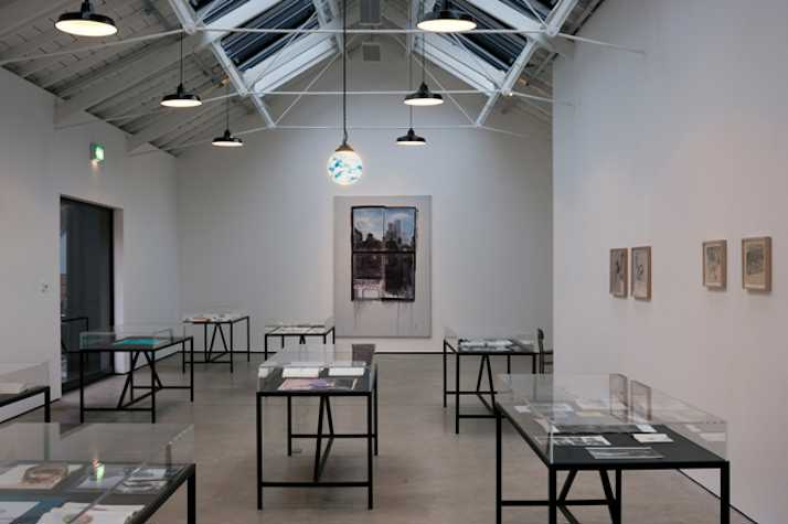 gallery space with multiple black metal tables encased by glass tops. they contain notebooks. on the back wall there is a large canvas with a painting of a view from a window. on the right wall there are four framed drawings.