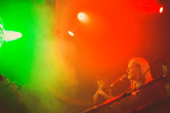 In a dark room lit in different colours, there is smoke, and a woman speaking into a microphone while looking into a piece of paper. On there side there is a woman standing and taking a picture of her with her phone and a photographer taking a picture of her, in front of her, with a DSLR camera.