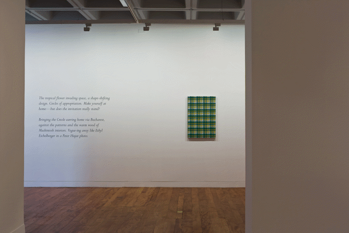 Black cursive text on a gallery wall that begins 'The tropical flower, invading space. A shapeshifting design'. A canvas of tartan fabric hung on the gallery wall.