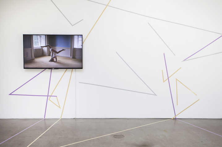 A white wall with painted colorful straight lines intersecting. There is a screen showing a video of someone doing a handstand.