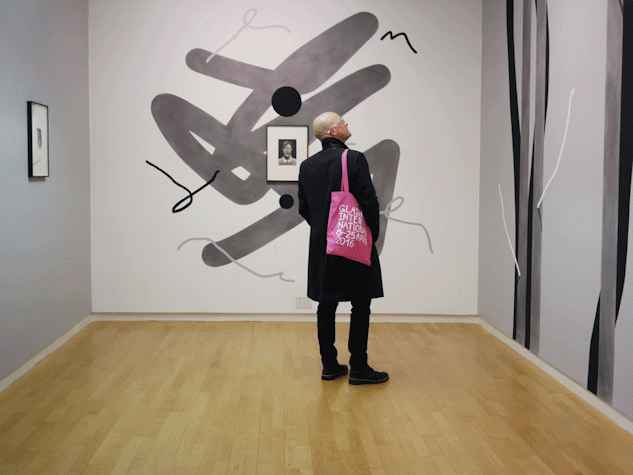A person wearing black with a pink bag in an exhibition space. The walls are painted with black and grey shapes. Two framed works are visible on the walls.