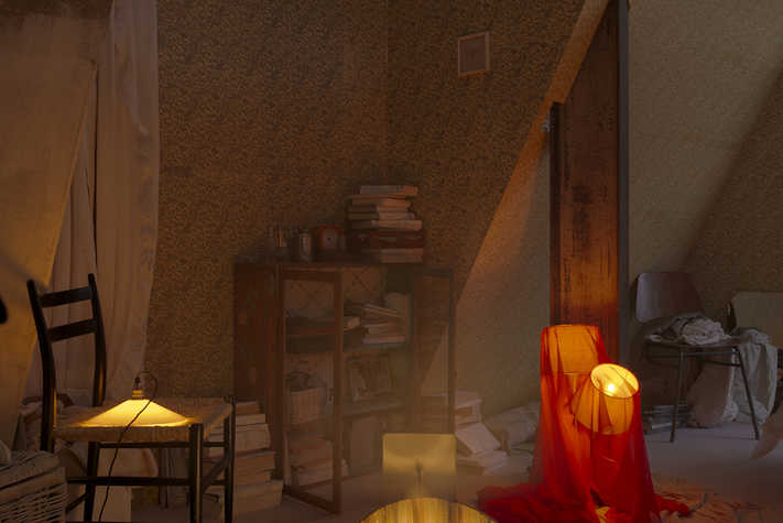 In a dimly lit room, there is furniture including a chair. a bookcase stacked with books, and on the floor there is a lamp covered in a thin fabric.
