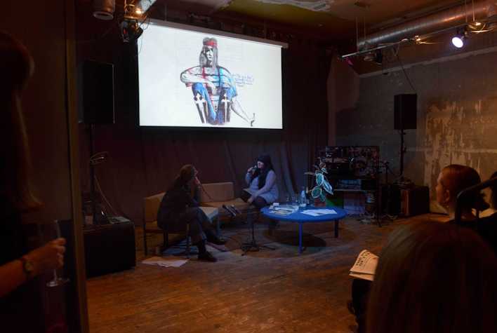 In a dimly-lighted room Fiona Jardine is in conversation with Claire Biddles with a proyection of a drawing on top of them. There is an obscured audience in front of them.