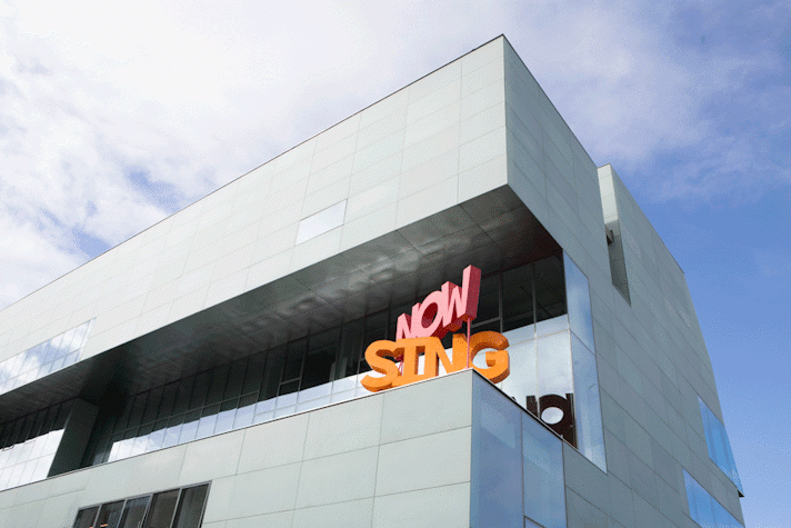 The words "Now Sing" are standing on stands, as a bold block sculpture. They are bright coloured. They are set on the terrace of a modern building.