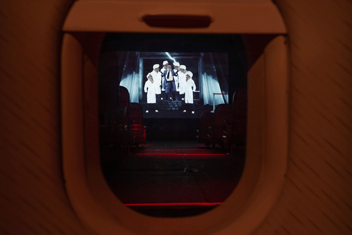 The image shows Jenkin van Zyl's installation at Tramway. Framed by an aeroplane window is a projection screen showing strange characters in lab coats.