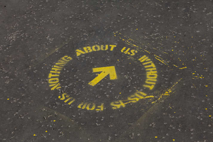 yellow spray paint stencilled onto asphalt floor. it reads 'nothing about us without us is for us' in a circle with an arrow in the middle