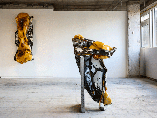 A room with concrete floors and columns has a white wall with a orange, black and whitesculpture made of metal. There is another in front of it, made of the same materials.