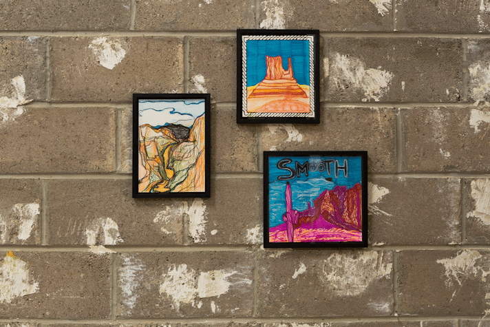 Three black framed felt-tip pen drawings hang on a concrete block work wall. They show landscapes in crude blue, orange and pink. In one, the landscape is pink and sky is blue and the word 'smooth' floats in the sky in bubble writing above a pink cactus.