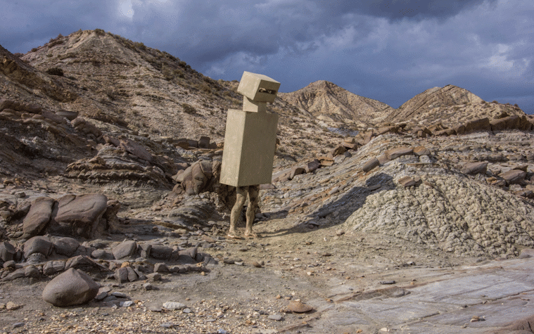 Film still showing a rocky landscape and a figure in box-shaped costume