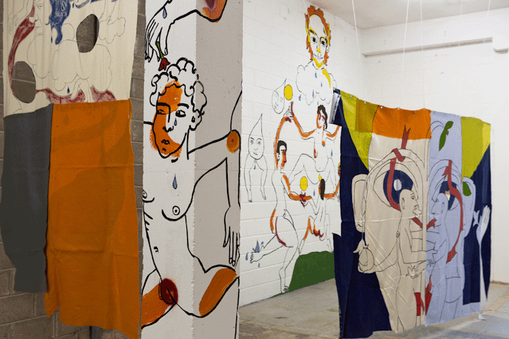 Playful and surreal line drawings of figures with blocks of colour cover a wall, column and fabric hanging in an industrial space