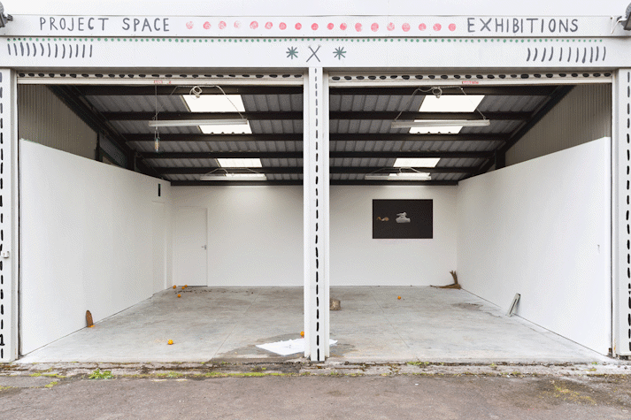 objects including a white towel and oranges are strewn across a concrete floor space. One side of the space is open to the outside. In the background a dark print is pasted to the wall