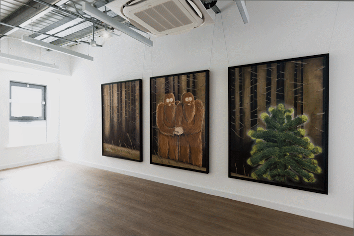 three large scale pastel drawings in black frames and portrait orientation hang on a white wall. they have a shared backdrop of a dark leafless forest. the left drawing shows tree trunks, the centre drawing shows two yeti figures embracing one another, the right drawing shows a monkeypuzzle tree.