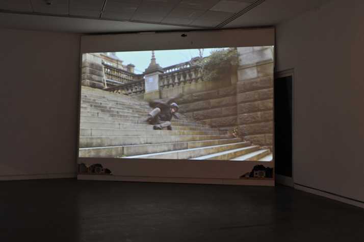large projection screen in the corner of a white empty room. image on the screen is a man falling down outdoor steps