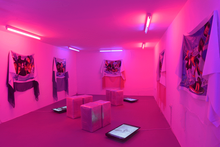 A room with pink neon lights has three screens on the floor, next to sitting blocks and on the walls it has five fabric hanging with images of women on it.