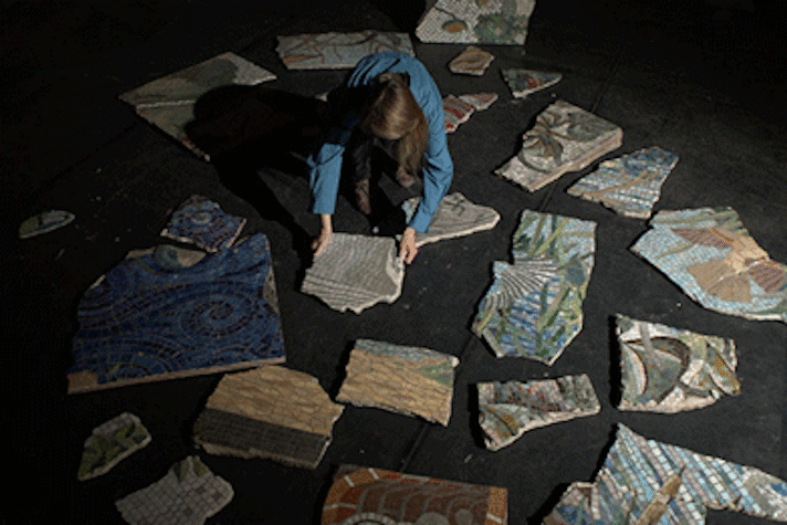 Film still, person kneeling on floor arranging pieces ofceramic artwork