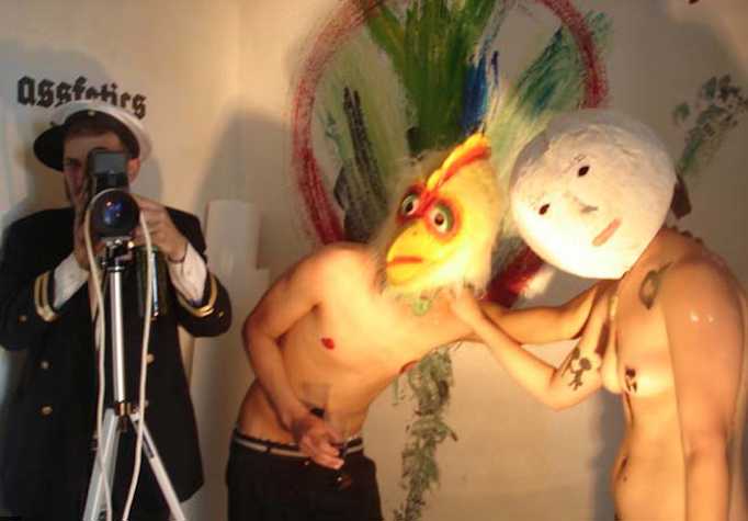 three performers. one is dressed in naval uniform. and the other two are topless, one with a chicken mask and the other white a white round mask with a face.