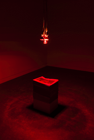 nstallation view, drop lights from ceiling illuminating the space red and a rectangular plinth with papers on top