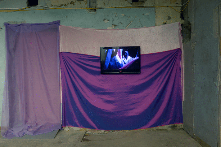 A room with several fabrics of purple colours hung on the wall and a tv screen with a video as a part of E. Jane's installation in Kelvin Hall as part of Glasgow International 2018.