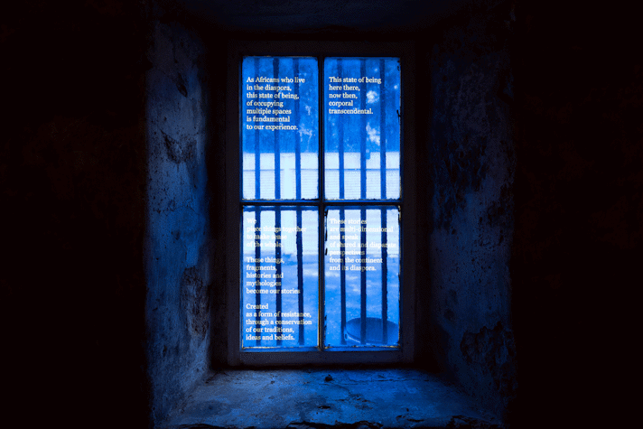 A blue film covers a window split into four panes, there is clear text on the film which begins 'As Africans who live in the diaspora, the state of being, of occupying multiple spaces is fundamental to our experience'.
