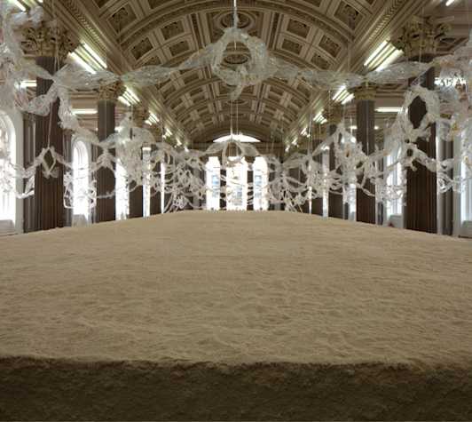 sculptural works hanging from ceiling in an ornate hall, with a large pile of sand on the ground