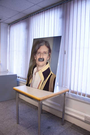 portrait photo of a person with a grey moustache, on a table leaned up against a window with white blinds.