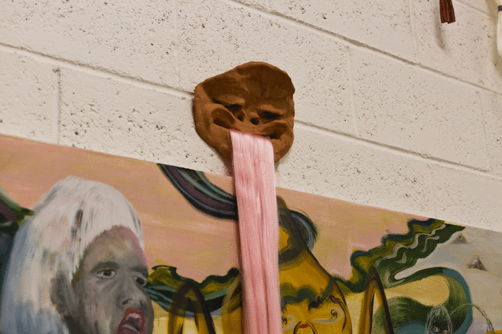 detail of clay mask hanging on a white blockwork wall, pink hair falls from the mouth.