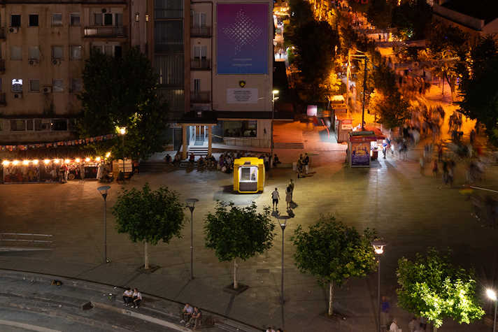 A yellow pod sits in the middle of a city setting, at night time, with people milling around.