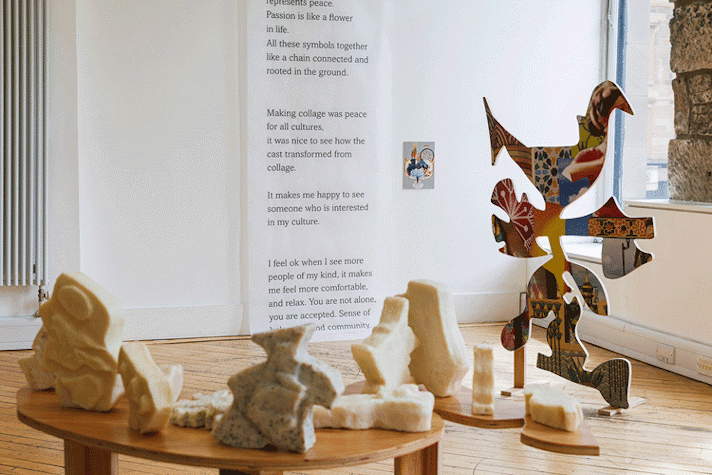 Sculptures are displayed on a wooden table and the floor, with artwork featuring written text on a wall behind.